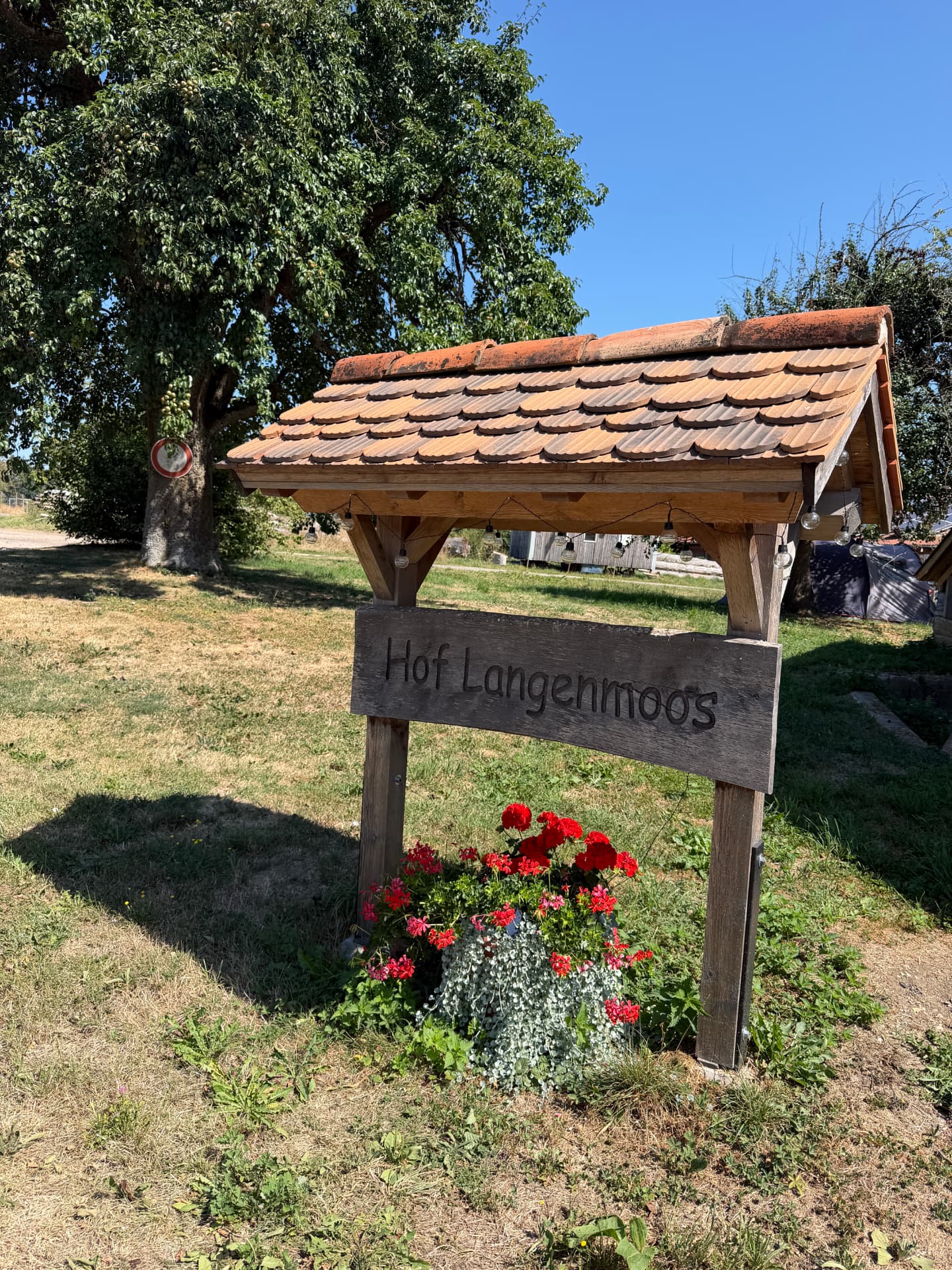 Holzschild „Hof Langenmoos“ unter einem kleinen Ziegeldach am Hofeingang